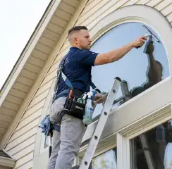 Professional window cleaner on a ladder using a squeegee to clean an exterior arched window, Anchorage, AK.